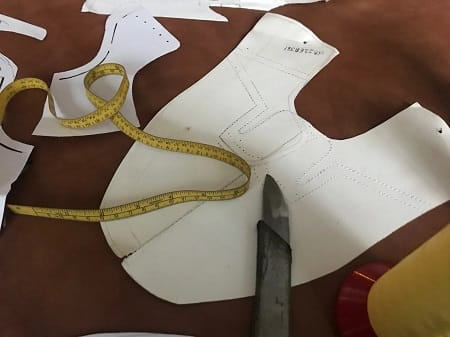 Cartamodello di una tomaia con metro da sarto e trincetto, appoggiato su una pelle marrone